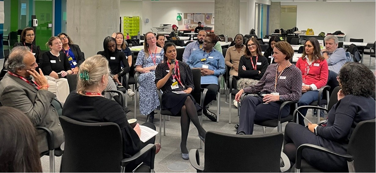 Two women sitting two chairs, while other people sit around them. One of the women has a microphone and is talking. Everyone listens and participates in the workshop